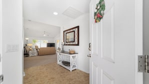 A hallway leading to a living room with a white door, a console table with a mirror, and a beige carpet. - Pet-Perfect Village Retreat with Fenced Yard in a Prime Spot – Book & Save (The Villages)