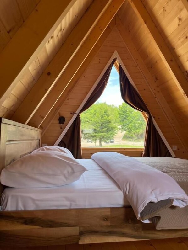Un dormitorio acogedor en el ático, con techo inclinado de madera, una cama individual con ropa blanca y una ventana con vistas a un paisaje verde.