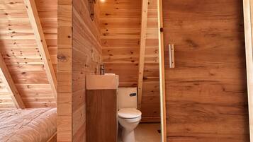 A wooden cabin interior with a loft bed, a wooden bathroom with a toilet, and a small sink.