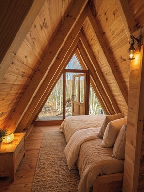 Interior de madera con techo inclinado, una cama con almohadas, una mesita de noche con lámpara y una ventana con vista a árboles.