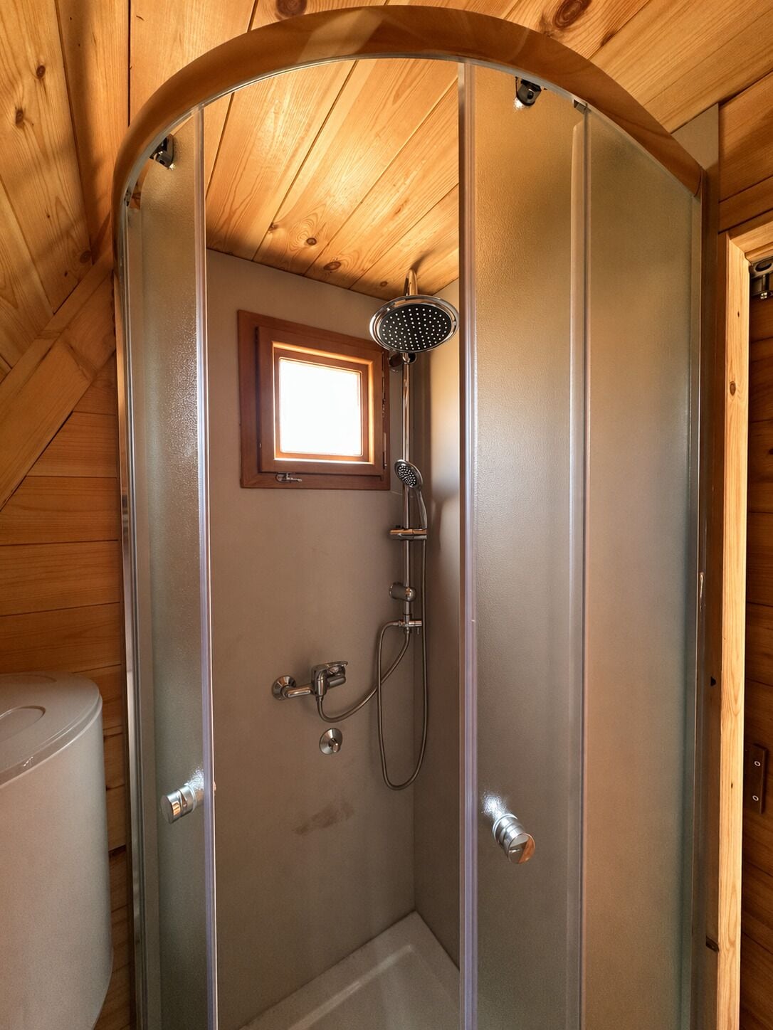 A wooden-paneled bathroom with a glass shower enclosure, a window, and a toilet.
