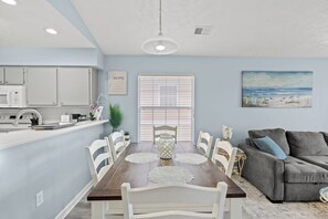 A dining area with a wooden table, white chairs, a gray sofa, and a painting of a beach scene. - Surf & Sail | cozy with TONS of amenities (Myrtle Beach)