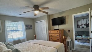 Une chambre à coucher comprenant un lit, un ventilateur de plafond, une commode, un téléviseur et une porte donnant sur une autre pièce.