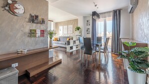 Un salon moderne avec un plancher en bois, un canapé, une table à manger avec des chaises, un coin cuisine et une grande fenêtre avec des rideaux.