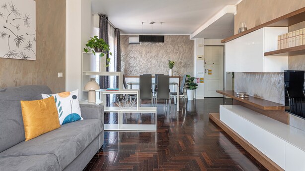 Un salon moderne avec un canapé gris, une table basse et un meuble pour la télévision. On y trouve également un coin repas avec une table et des chaises, et une cuisine à l’arrière-plan.