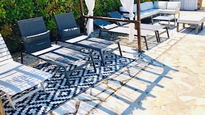 Een patio met buitenmeubilair, waaronder een loungeset en fauteuils, een patroonrijk vloerkleed en een pergola met witte gordijnen.