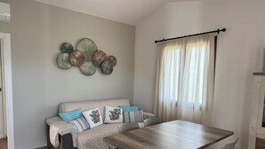 A dining area with a wooden table and white chairs, a beige sofa with patterned cushions, and a decorative wall piece above the sofa. - House surrounded by olive grove 900mt from the sea (Orosei)