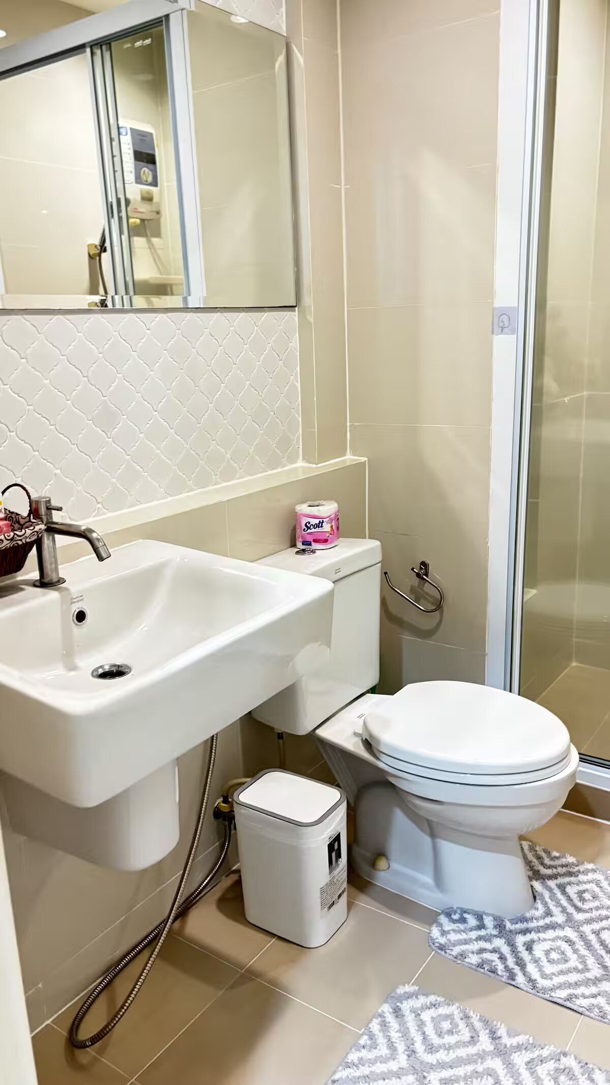 Une salle de bain avec un lavabo, des toilettes et une douche, le tout de couleur blanche. On trouve un rouleau de papier toilette sur le réservoir des toilettes et une petite poubelle à côté.