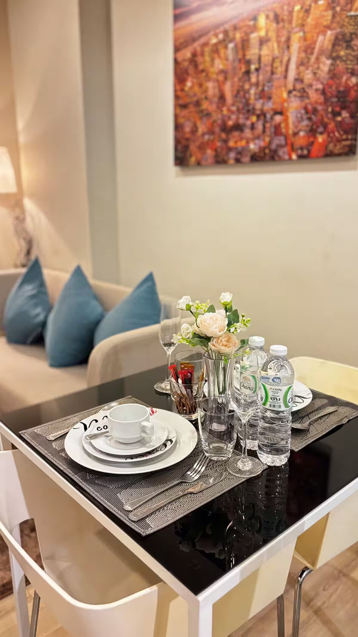 Una mesa de comedor dispuesta con platos, vasos, cubiertos y botellas de agua, con un jarrón de flores en el centro.