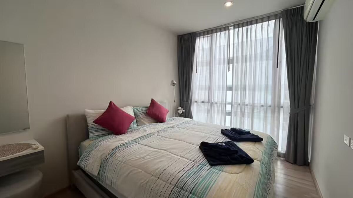 A neatly made bed with a striped bedspread and two pillows, a bedside table with a lamp, and a window with sheer curtains.