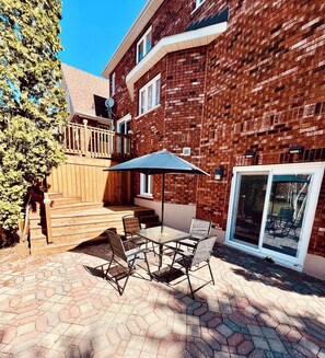 A patio with a table and chairs, a wooden deck, and a brick building with a glass door. - Cozy Basement Apartment in Timmins (Timmins)