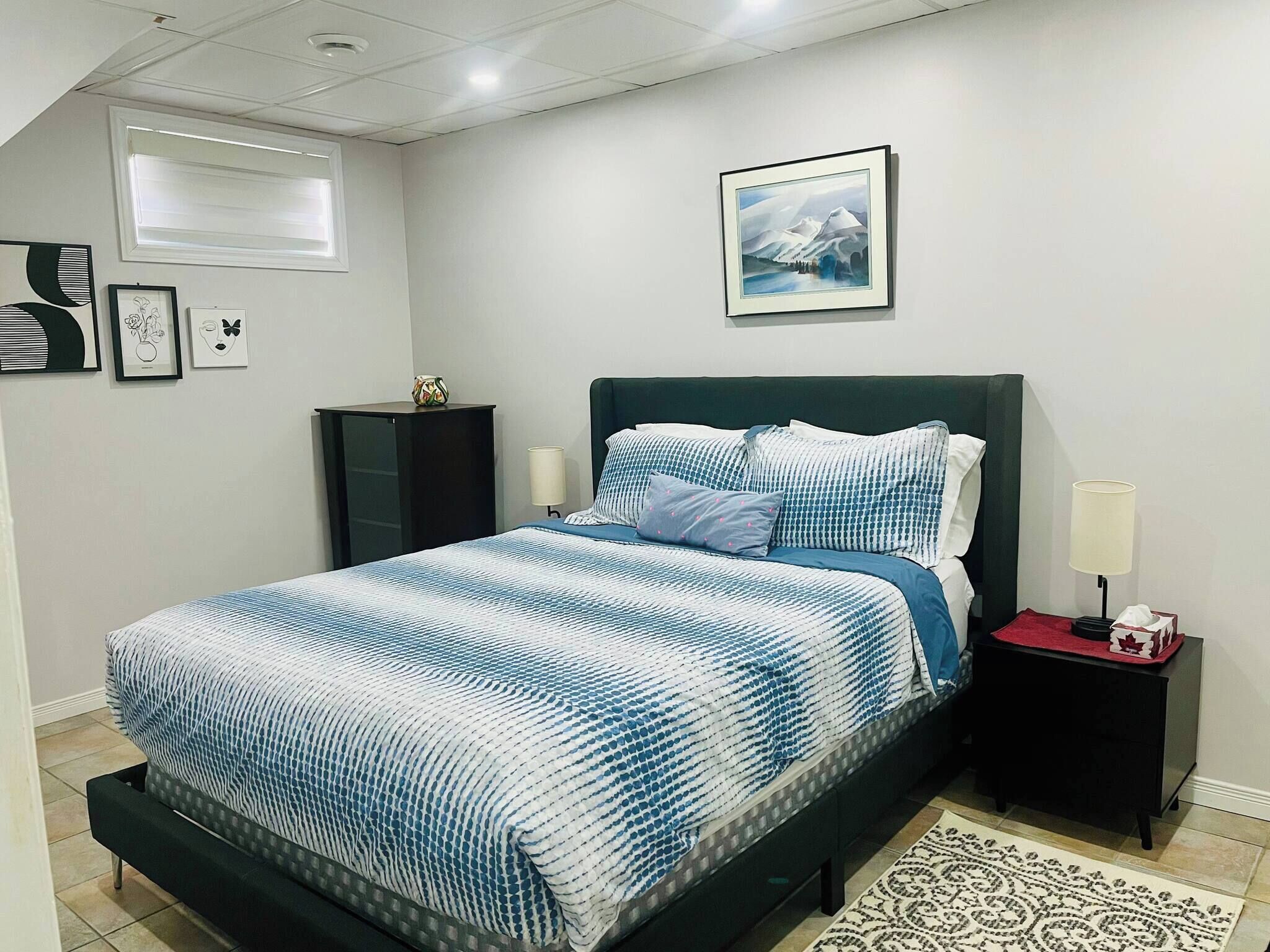 A bedroom with a bed, a nightstand, a lamp, a framed picture, and a window.