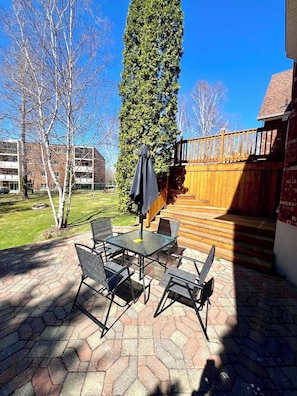 A patio with a table and chairs, a wooden deck, and a tall tree.