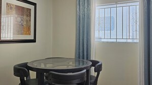 A small, well-lit room with a round glass table and four chairs. There is a framed artwork on the wall, a window with blinds, and a curtain. - Home sweet Home (Surrey)