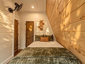 Een slaapkamer met een schuine kap, een bed met een groen en bruin dekbedovertrek, een houten deur en een wandlamp.