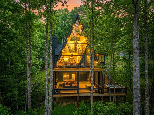 A modern, triangular wooden cabin with a glass facade, surrounded by dense forest. - Brand New • Epic Creek-Front A-Frame • HotTub • FirePit • Games • Creek Access (Epworth)