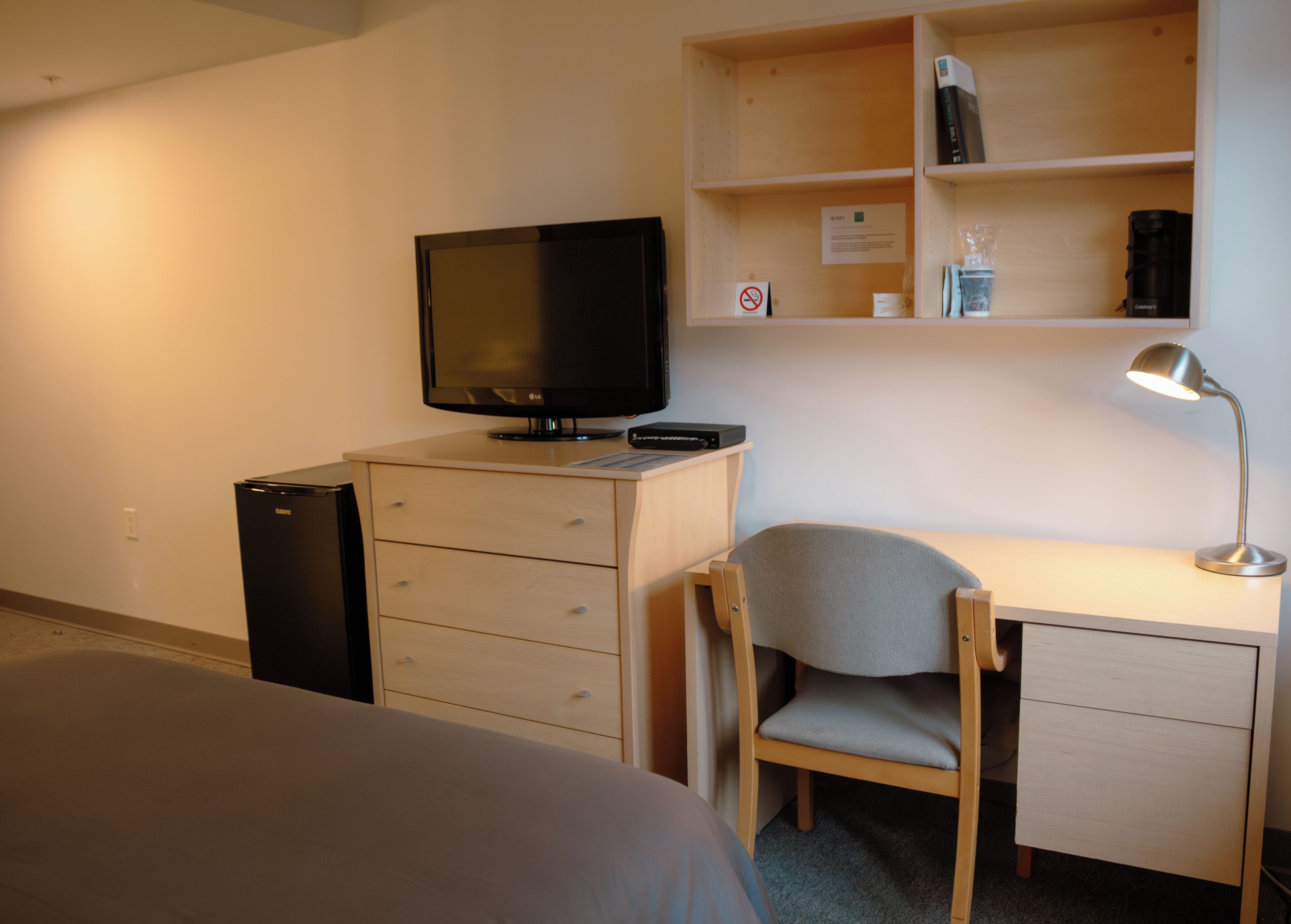 Habitación de hotel con cama, escritorio con silla, televisor, cafetera y estante con libros y un vaso.