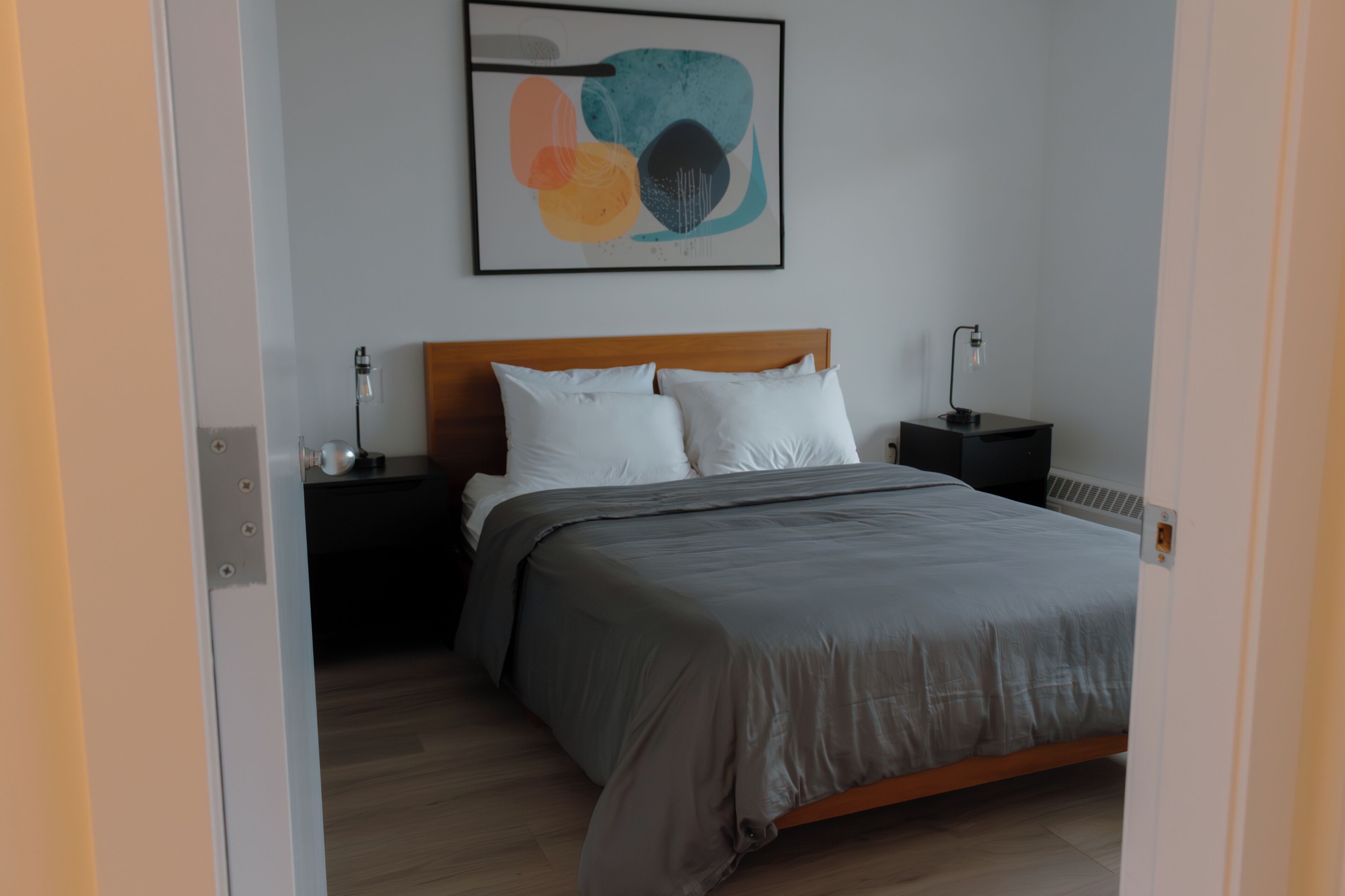 Quarto com cabeceira de madeira, colcha cinza, duas almofadas brancas, mesa de cabeceira com abajur e uma obra de arte abstrata na parede.