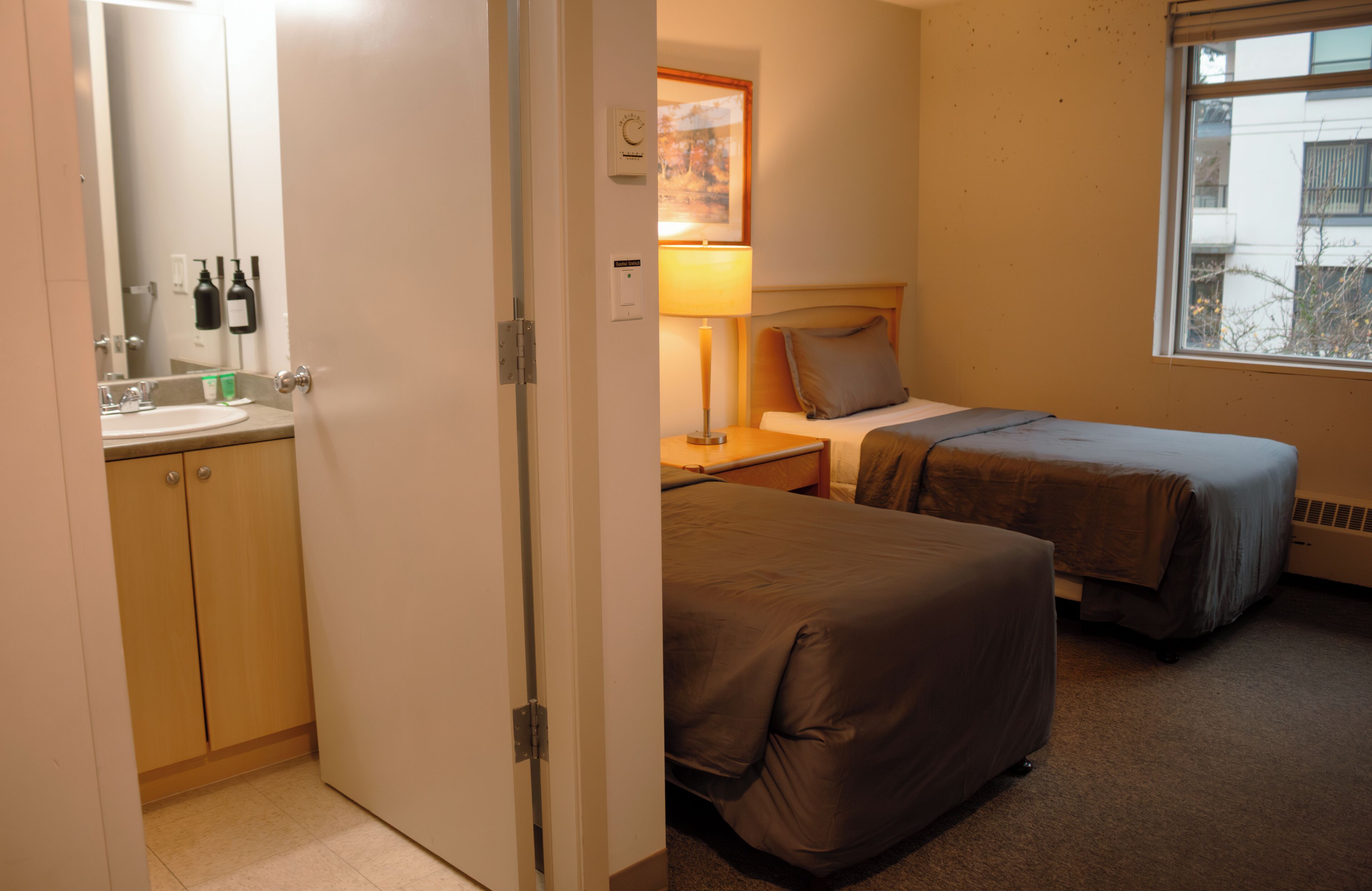 Habitació d'hotel amb llit, taula de nit, làmpada i finestra que mostra edificis.