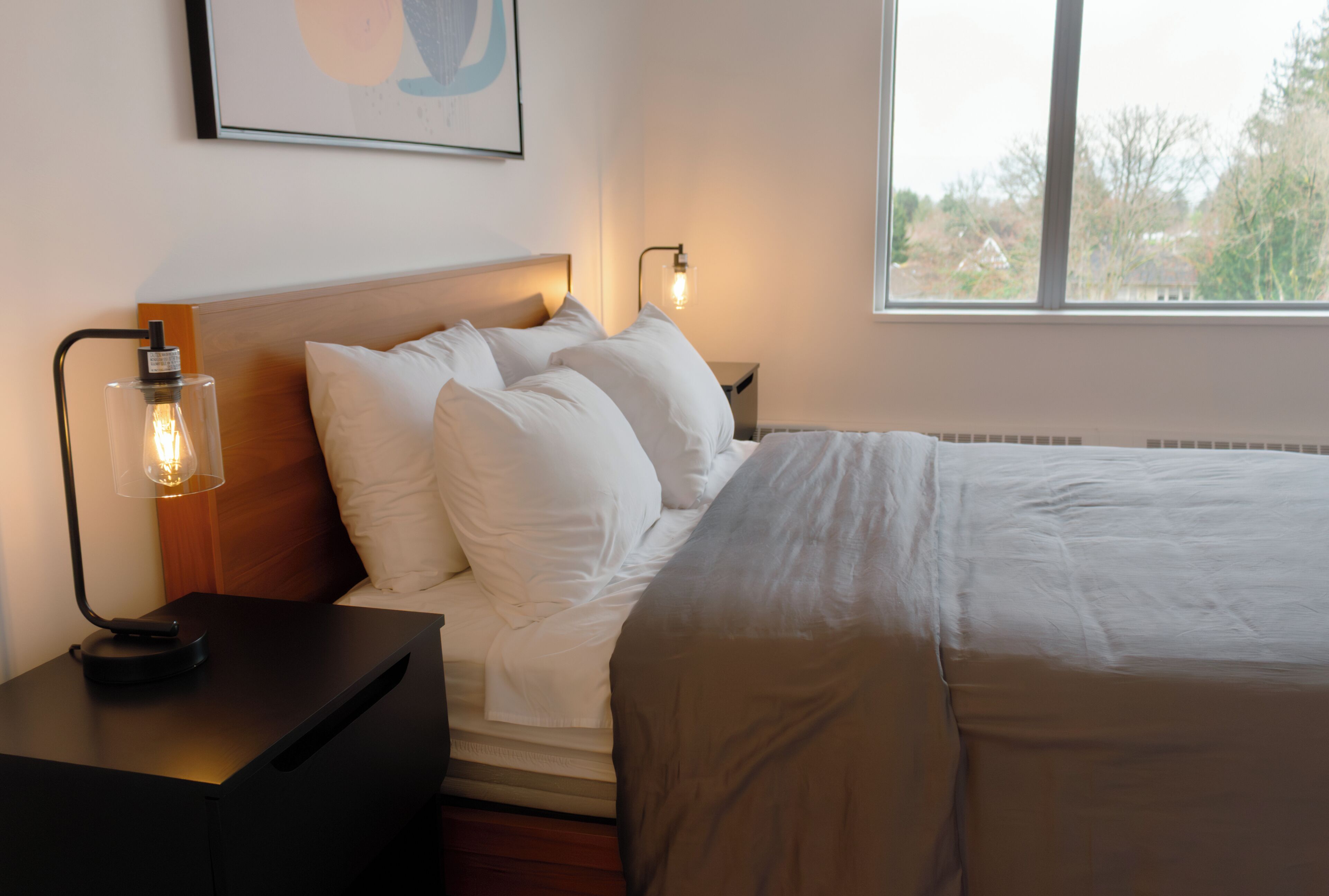 Een net opgemaakt bed met witte lakens, een bedlampje en een wandlamp.