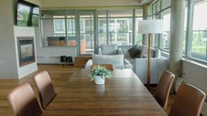 A modern living room with a wooden dining table, grey sofas, and a large window providing a view of the outdoors. - Carey Lodge at UBC (Vancouver)
