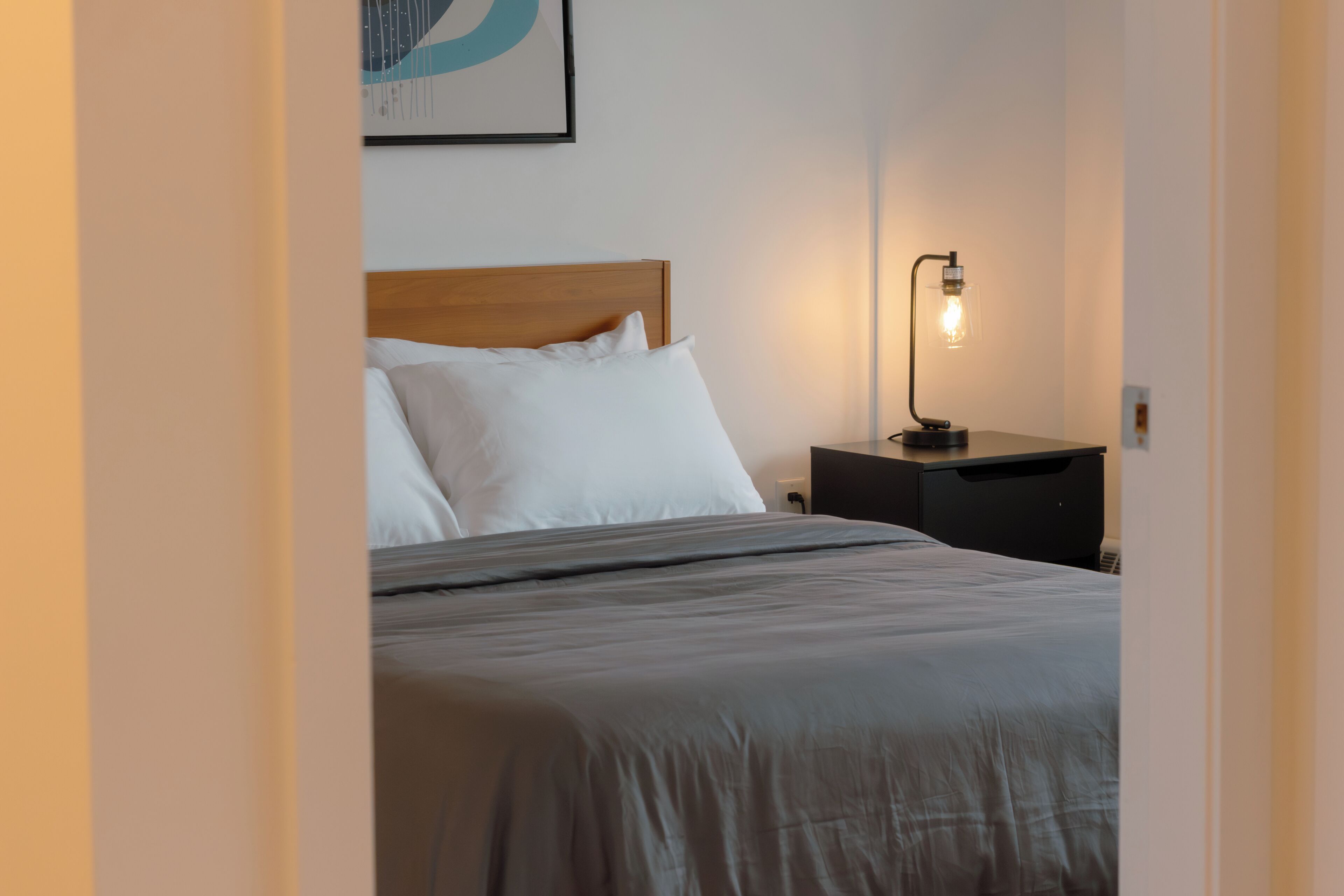 Una cama bien hecha con ropa de cama blanca y gris, un cabecero de madera y una mesita de noche con una lámpara.