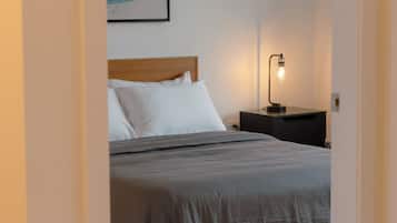 Una cama bien hecha con ropa de cama blanca y gris, un cabecero de madera y una mesita de noche con una lámpara.