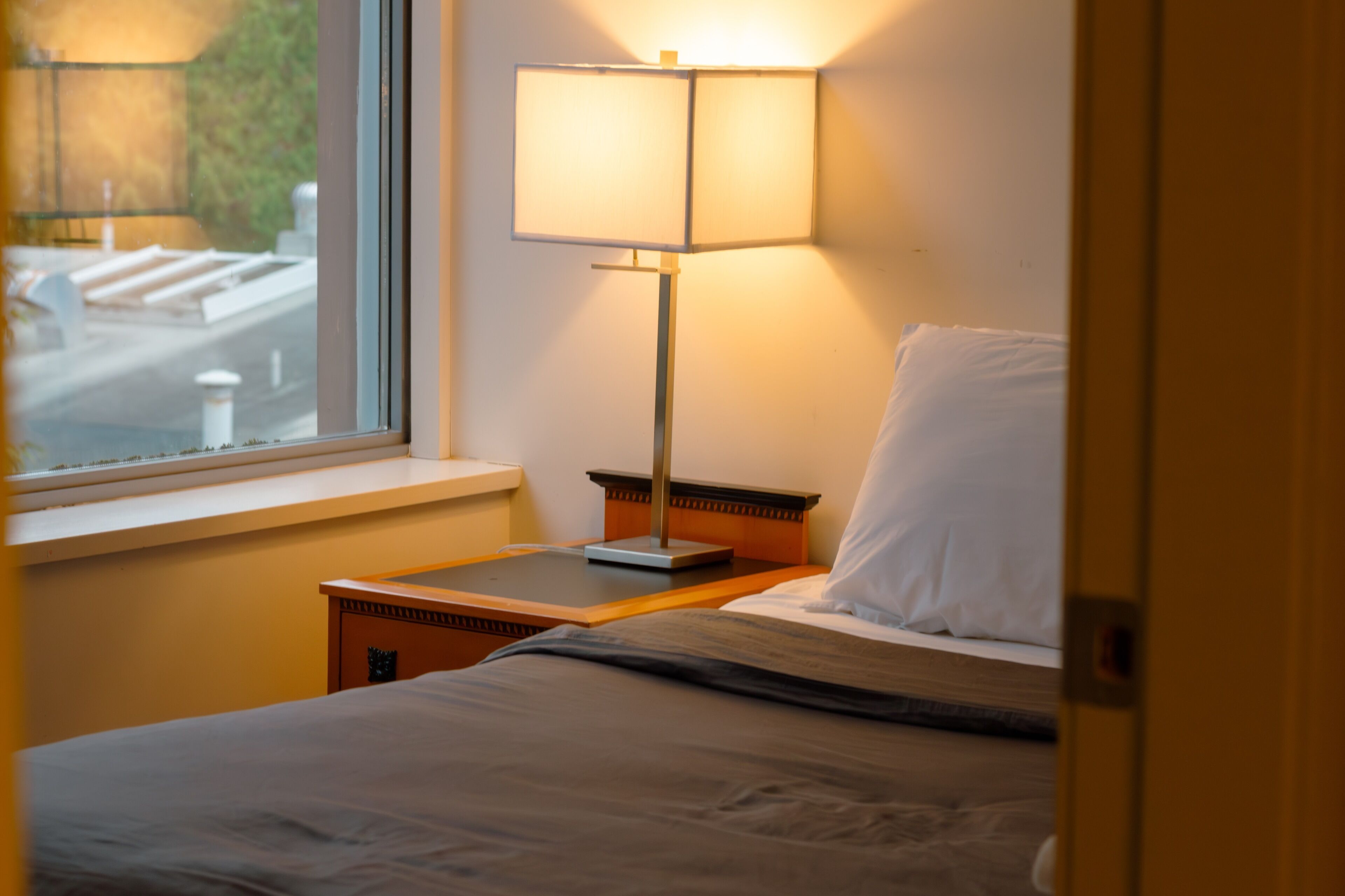 A hotel room with a bed, a nightstand, a lamp, and a window with a view of a street.