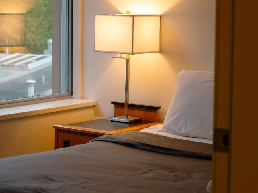 A hotel room with a bed, a nightstand, a lamp, and a window with a view of a street.