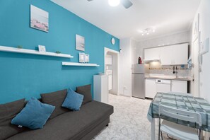 Ein Zimmer mit blauer Wand, einem grauen Sofa mit blauen Kissen, einem kleinen Tisch mit grüner Tischdecke und einer Küchenzeile mit weißen Schränken und Geräten.