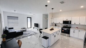 A modern kitchen with white cabinets, a central island, and stainless steel appliances. There is a dining area with a black table and chairs, and a living room with a black sofa and a TV. - Casa encanto, your home away from home. (Lehigh Acres)