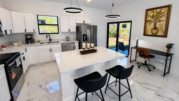 A modern kitchen with white cabinets, a central island with bar stools, a dining area with a table and chairs, and a large window offering a view of the outdoors. - Casa encanto, your home away from home. (Lehigh Acres)