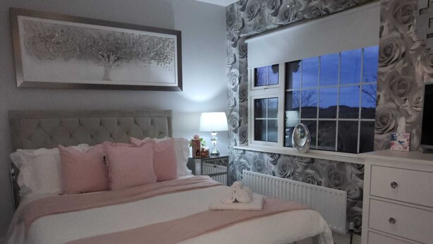 Un dormitorio con cabecera capitonada, cama con ropa de cama rosa, mesita de noche con lámpara y ventana con vistas al exterior.