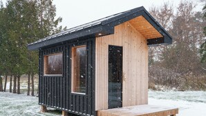 A small cabin with a wooden exterior and a black roof, featuring a large window and a door. - Cozy Collingwood Farmhouse, With Sauna (Collingwood)