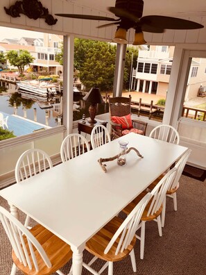Un espace repas avec une table et des chaises blanches, une branche décorative en guise de centre de table, et une vue sur un plan d’eau et des bâtiments.