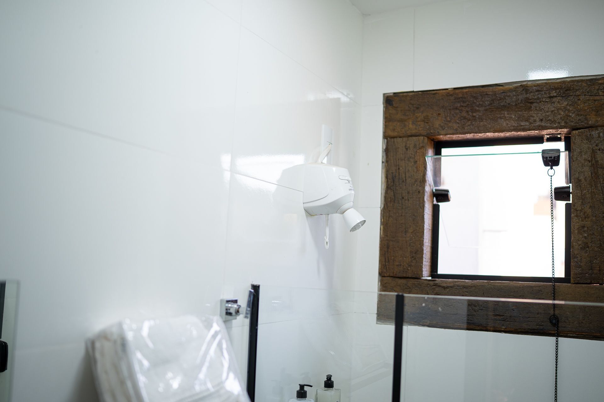 Een badkamer met witte tegels, een aan de muur bevestigde haardroger, een raam met houten kader en een douchegedeelte.