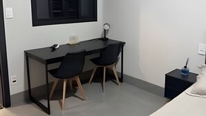 Una habitación pequeña y moderna con un escritorio, dos sillas, una ventana con persianas, un equipo de aire acondicionado y una mesita de noche con lámpara.