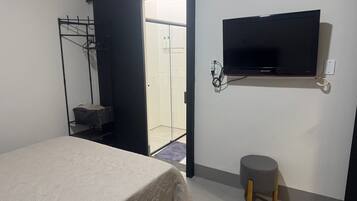 Quarto com uma cama, televisão fixada na parede, um banquinho pequeno e uma porta que dá para um banheiro.