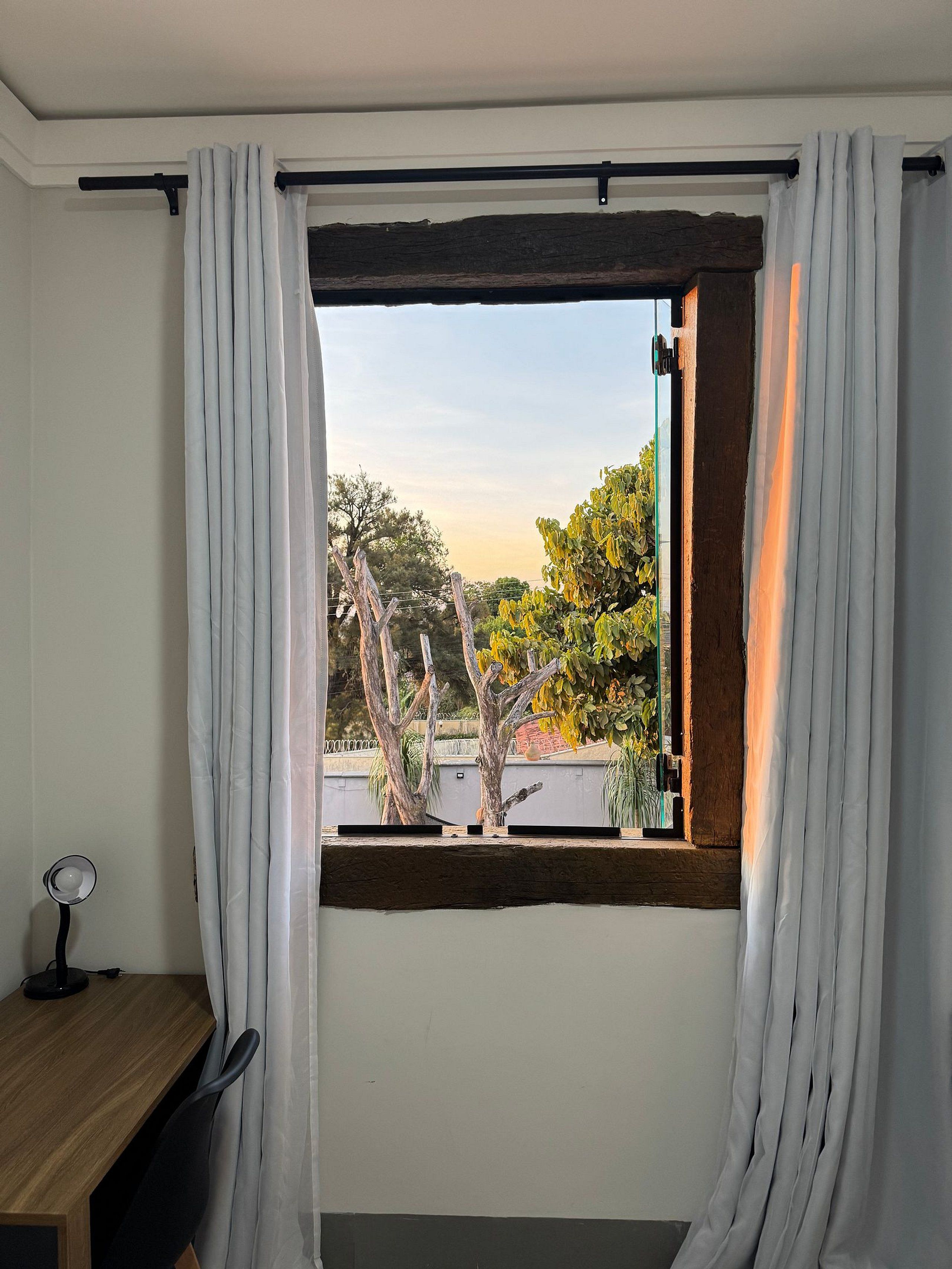 A room with a window, white curtains, a wooden desk, and a chair.
