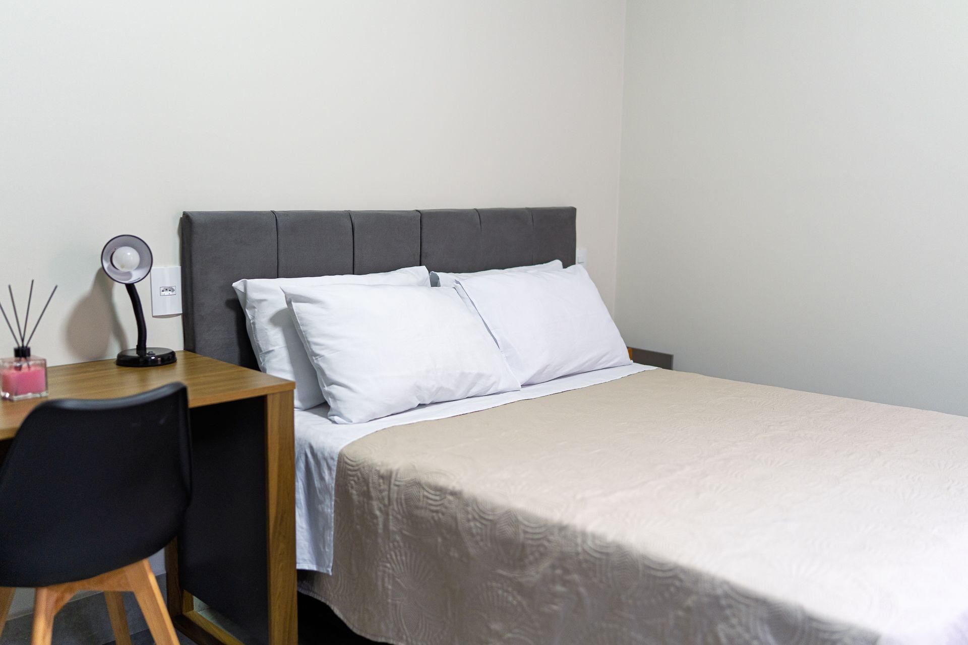 Una cama bien tendida con sábanas blancas, un cabecero tapizado en gris y un pequeño escritorio con una lámpara y una vela rosa.