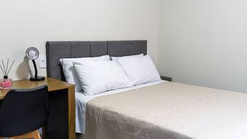 Una cama bien tendida con sábanas blancas, un cabecero tapizado en gris y un pequeño escritorio con una lámpara y una vela rosa.