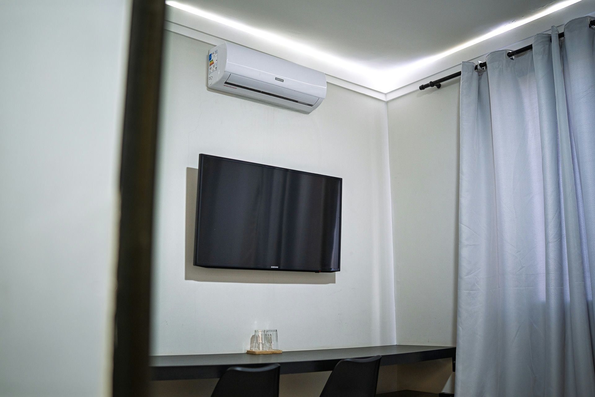 Una habitación con televisión de pantalla plana en la pared, una mesa con sillas y una cortina en la ventana.