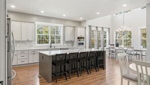 A modern kitchen with white cabinetry, a central island with bar stools, and a dining area with white tables and chairs. - Creekside Luxury Retreat on 22 Acres (Louisville)
