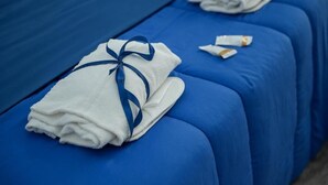 Hotellihuone, jossa on siniset sängyt ja siististi taitettu valkoinen pyyhe, jonka ympärille on sidottu sininen nauha.