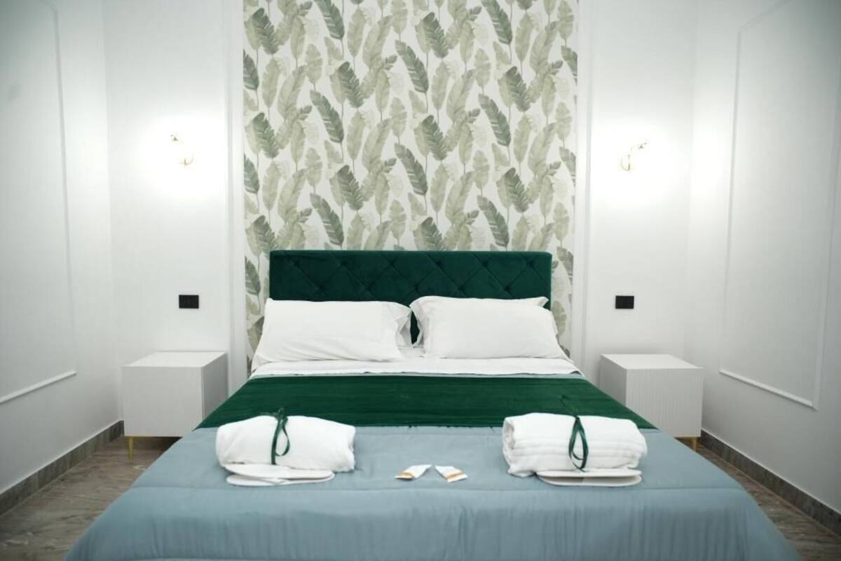 A neatly made bed with a green headboard, white pillows, and a blue bedspread. There are two bedside tables with lamps and towels on them.