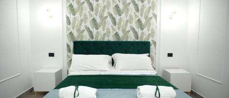 A neatly made bed with a green headboard, white pillows, and a blue bedspread. There are two bedside tables with lamps and towels on them.