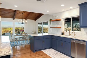 A modern kitchen with dark blue cabinets, a white backsplash, and a large window offering a view of the outdoors. - Harley’s Hideaway | Hilltop Views, Hot Tub & Pets (Paso Robles)