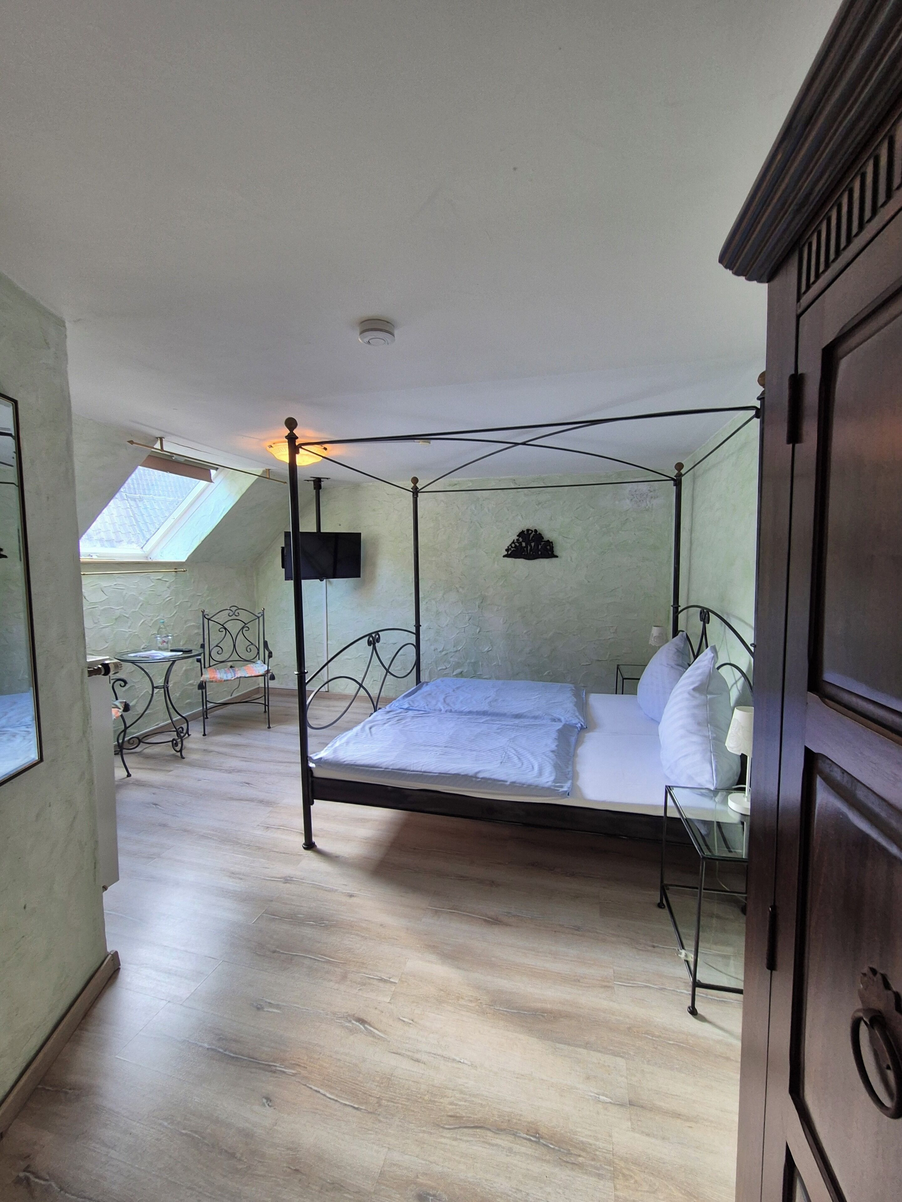 A bedroom with a four-poster bed, a skylight, a small table with a chair, and a television on the wall.