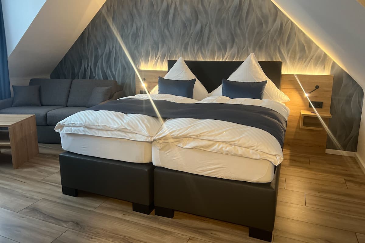 Une chambre de style loft moderne, avec un plafond en pente, un grand lit avec une literie blanche et foncée, un canapé gris et une table de chevet en bois avec une lampe.