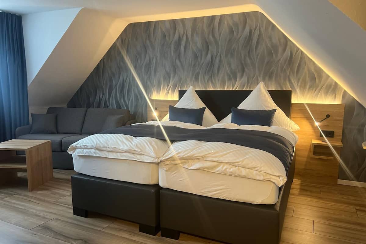 Une chambre à coucher moderne avec un plafond en pente, un lit avec une literie blanche et foncée, un canapé gris et un plancher en bois.
